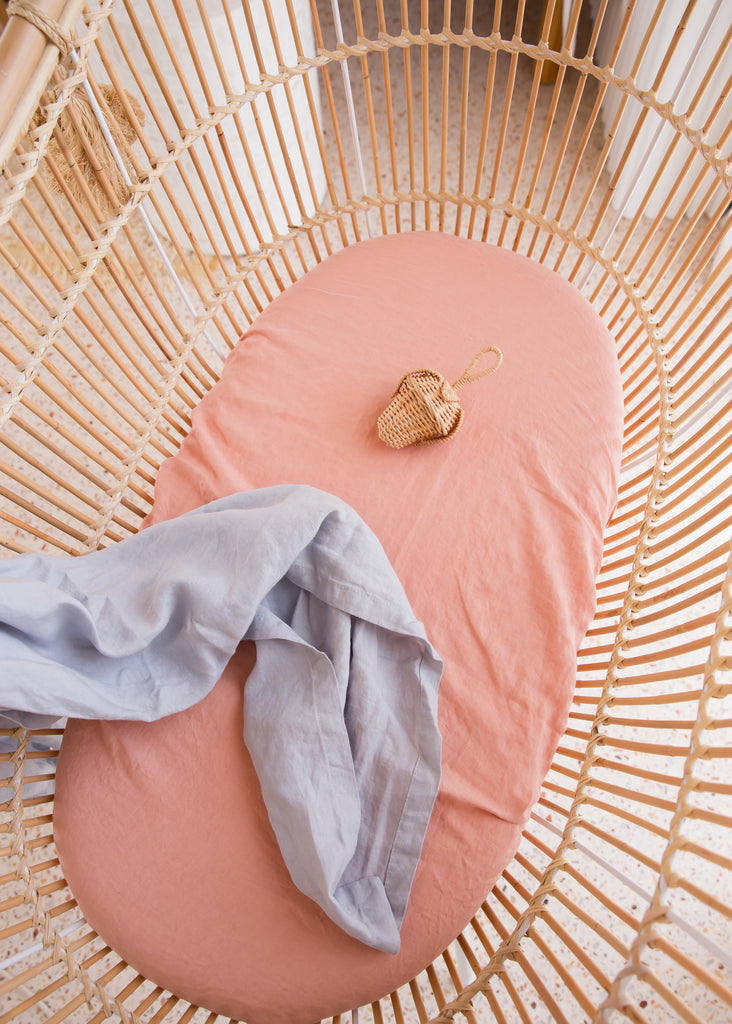 Coral Linen Bassinet Sheet with a Mist blue Linen Swaddle draped over the side of the bassinet